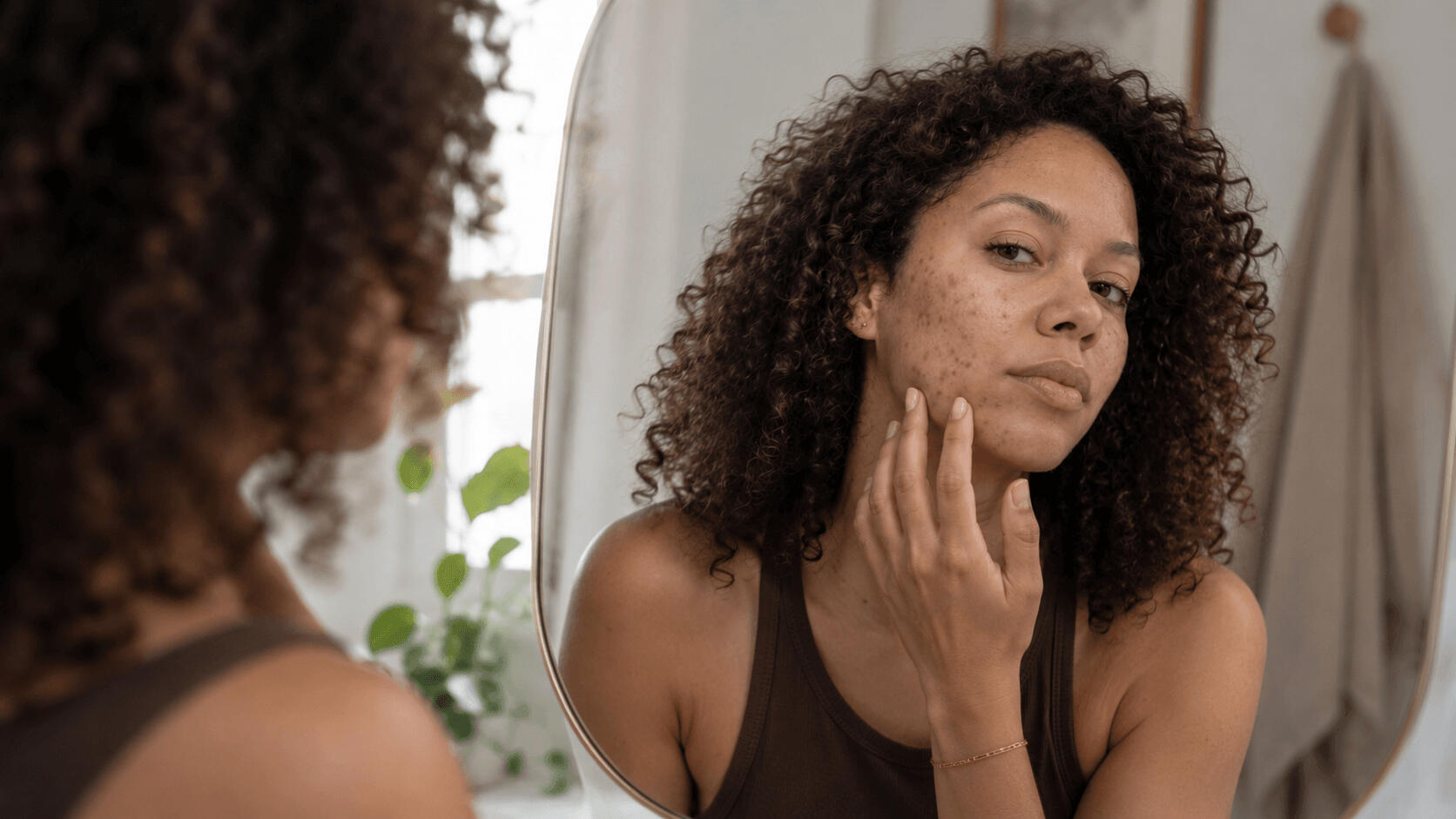 Adult woman in her 30s looking at her reflection in a bathroom mirror, examining recurring breakouts on her jawline that return in the same spot every month